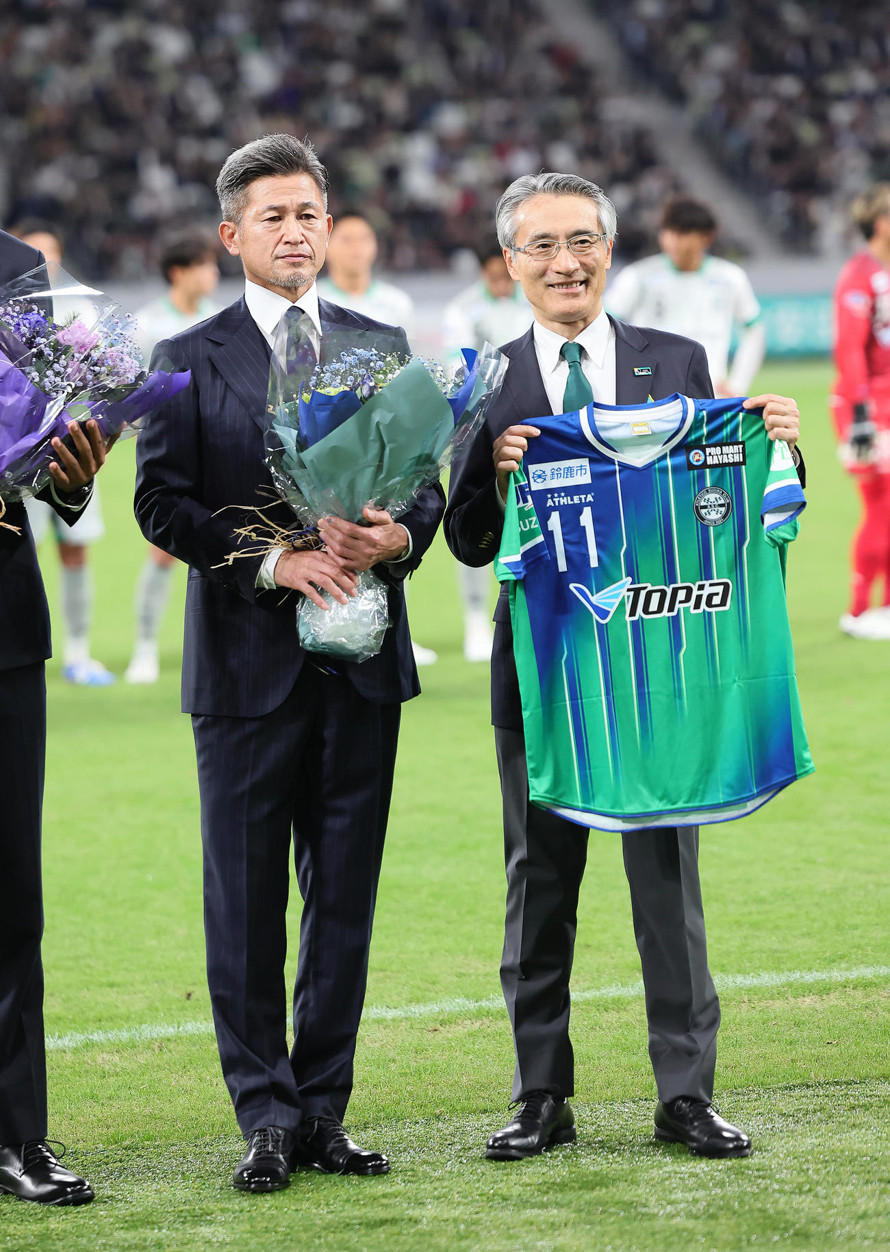 試合前のセレモニーで明治安田生命保険相互会社永島英器取締役代表執行役社長（右）から花束を受け取り記念撮影する鈴鹿カズ（撮影・足立雅史）