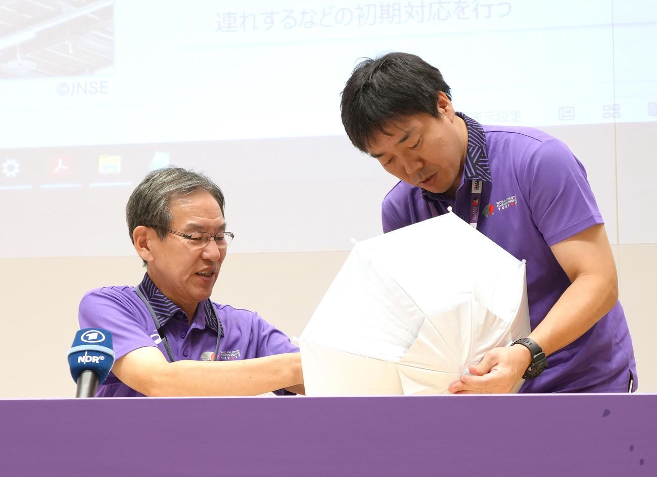 観客らに貸し出しするかさを頭につけ実演する東京2025世界陸上財団の武市事務総長（左）（撮影・足立雅史）