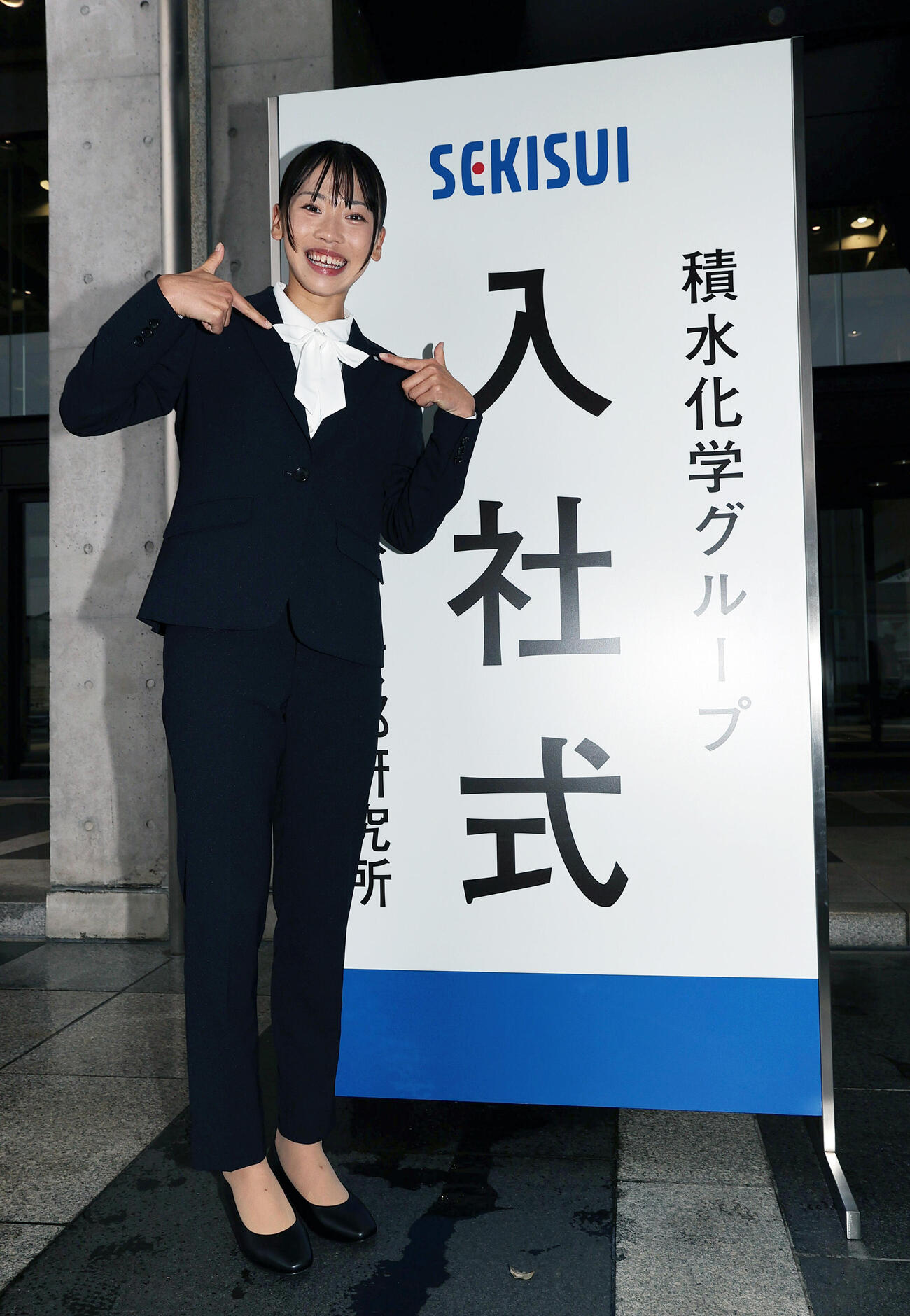 入社式を終えた積水化学の久保凛は胸元のリボンを指さし笑顔で見せる（撮影・上山淳一）