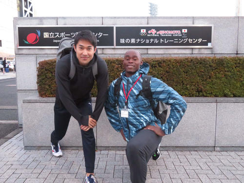 ナショナルトレーニングセンターの門の前で笑顔の飯塚（左）とシブシソ・マツェンジワ