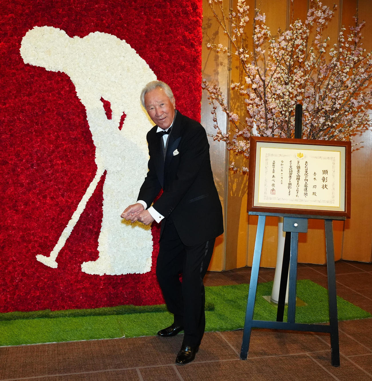 文化功労者顕彰を祝う会で展示されるカーネーションの花でつくられたモニュメントと同じポーズを見せる青木（撮影・江口和貴）