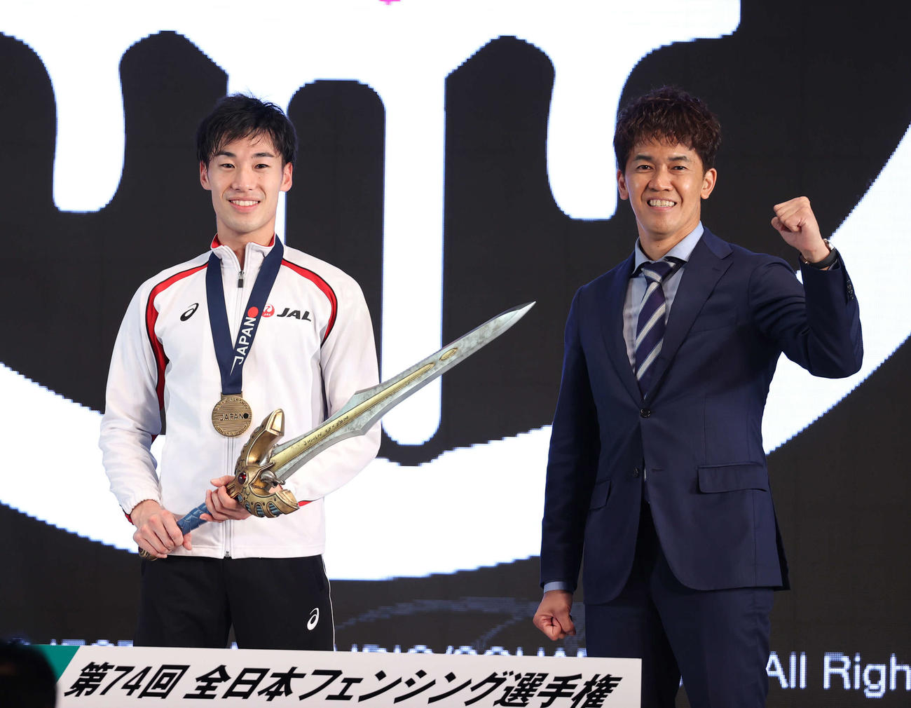 男子エペで優勝した加納（左）は協賛のドラゴンクエストウォークから提供された副賞の「ロトの剣」を手に笑顔を見せる。右は武井会長（撮影・河野匠）