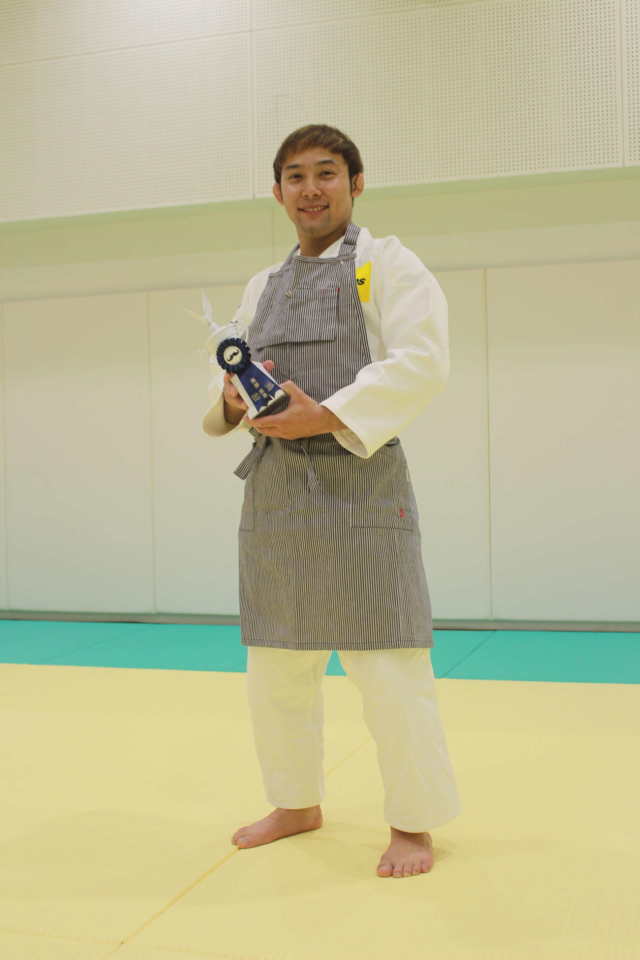 柔道着に副賞のエプロン姿の高藤はトロフィーを手に笑顔
