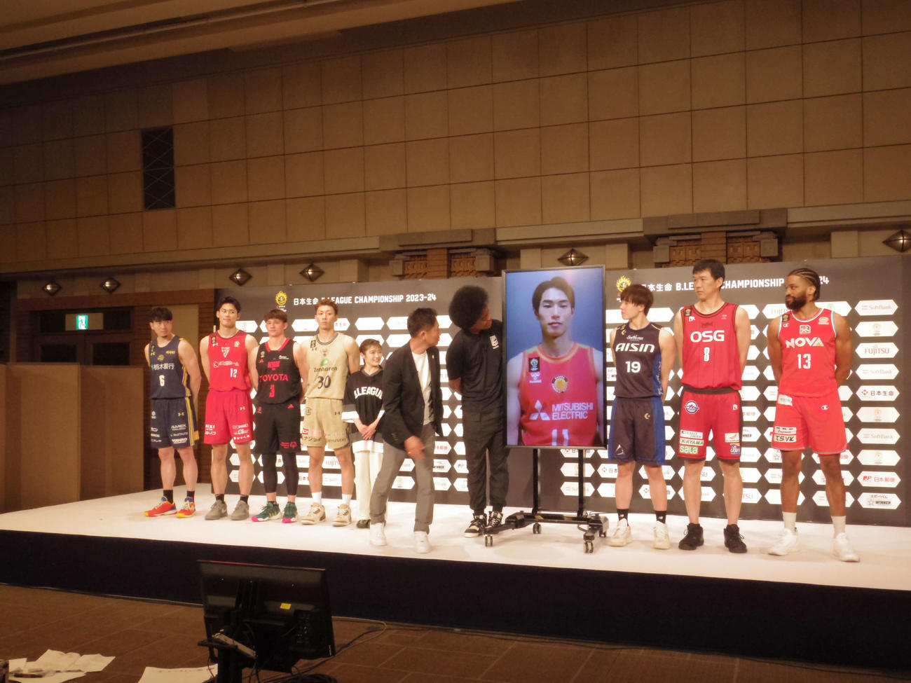 Bリーグのチャンピオンシップ進出会見で、ビデオ出演の須田侑太郎をのぞき込む島田チェアマン（左から6人目）やタレントの井上咲楽（左から5人目）、副島淳（左から7人目）や選手たち