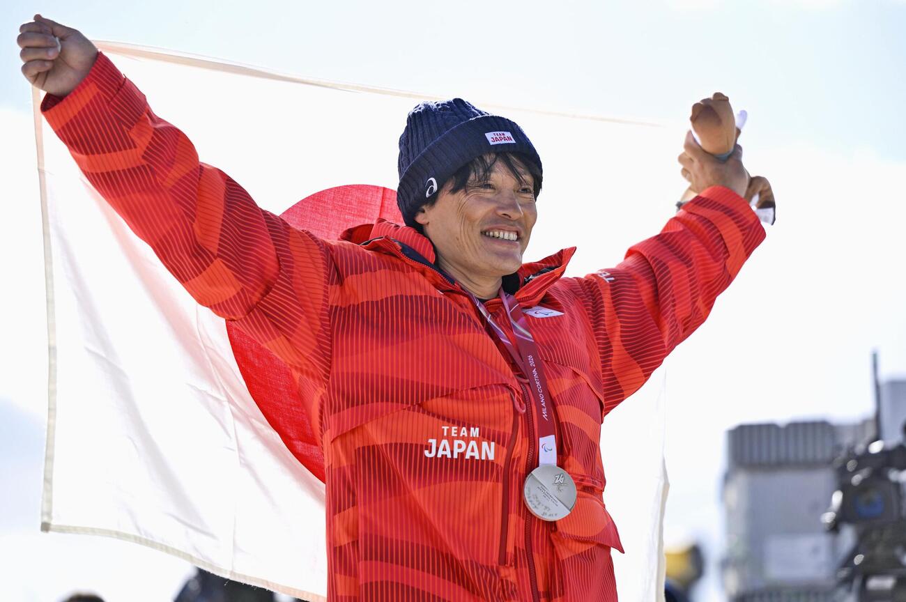 ミラノ・コルティナ冬季パラリンピックのスノーボード男子バンクドスラローム大腿障害で獲得した銀メダルを胸に、日の丸を掲げる小栗大地。冬季パラの日本選手団通算100個目のメダルとなった＝13日、イタリア・コルティナダンペッツォ（共同）