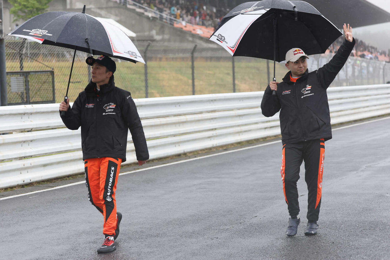 スーパーフォーミュラ第3戦　強い雨脚と霧が立ちこめる中で中止が決定し、コースに出てファンに手を振って歩く野尻智紀（左）と岩佐歩夢（JRP提供）＝2026年4月26日、大分県日田市のオートポリス
