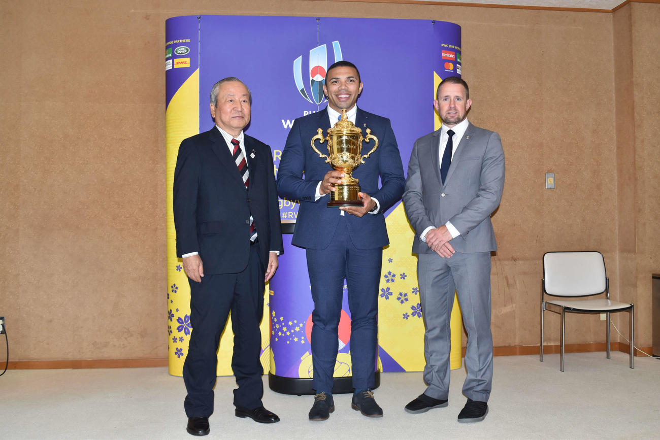 ラグビーW杯の優勝杯「ウェブ・エリス・カップ」を持つ、元南アフリカ代表WTBのブライアン・ハバナ氏。左はW杯組織委員会の嶋津昭事務総長。右は元ウェールズ代表WTBのシェーン・ウィリアムス氏